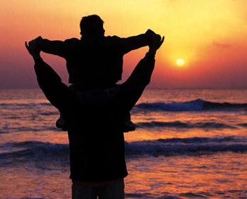 Son sitting on his father's shoulders, as they look at a sunrise across the ocean.