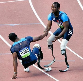 A runner in a race, with artificial legs, stops to help another runner up who has fallen.