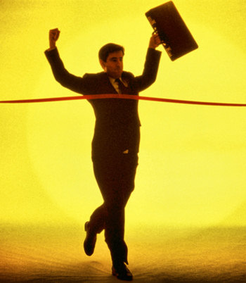 Picture of a man running with a briefcase, arms held high, breaking the red winners tape.