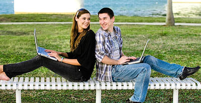 Young man and woman, sitting back-to-back on a park bench, holding computers, and smiling at each other.