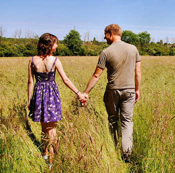 A young man and woman looking lovingly at each other as they're holding hands, while strolling through a large field together.